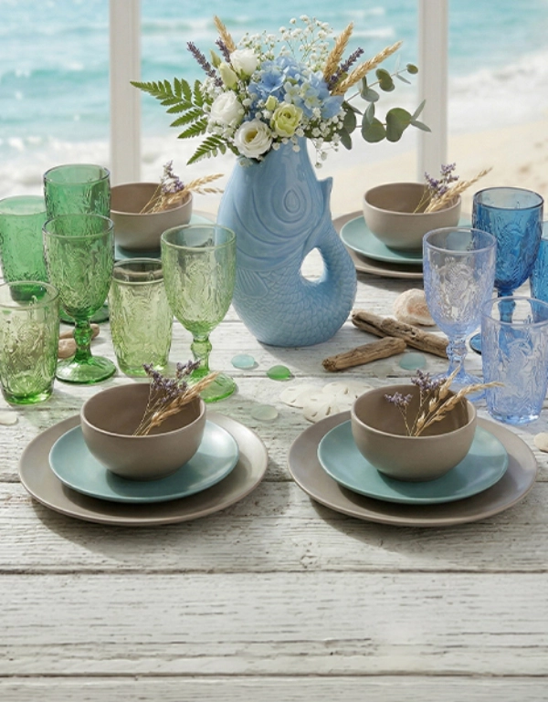 Stół udekorowany w stylu marynistycznym z kolorowymi kieliszkami, zastawą i wazonem z kwiatami na tle morza – stylowa dekoracja stołu na letnie chwile