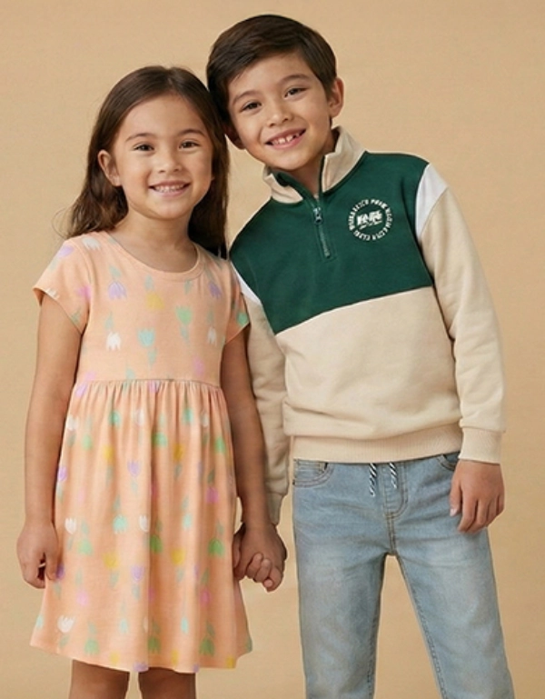 Dwoje dzieci stoi razem na tle jednolitego beżowego tła. Dziewczynka ma na sobie kolorową sukienkę, a chłopiec zielono-kremową bluzę.