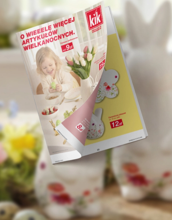 Dekoracyjna wiosenna scenka z trzema białymi figurkami królików, kolorowymi kwiatami i ulotką promującą produkty wielkanocne.