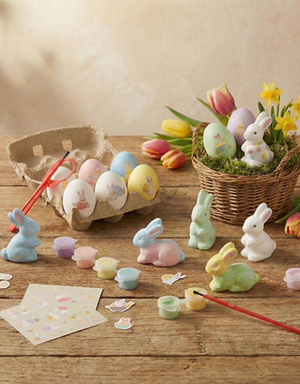 Kolorowe dekoracje wielkanocne z pomalowanymi jajkami, pastelowymi króliczkami i przyborami artystycznymi na rustykalnym drewnianym stole z kwiatami.