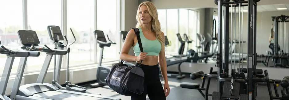 Osoba w stroju sportowym stoi na siłowni z urządzeniami treningowymi i trzyma czarną torbę sportową z logo Bench