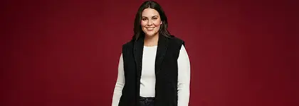 Osoba stoi pewnie przed jasnoczerwonym tłem, ubrana w białą koszulkę z długim rękawem i czarną kamizelkę. Osoba stoi pewnie przed jasnoczerwonym tłem, ubrana w białą koszulkę z długim rękawem i czarną kamizelkę.