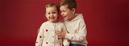 Dwoje dzieci pozuje blisko siebie, jedno w przytulnym swetrze w serduszka, a drugie w jasnej bluzie z kapturem, na czerwonym tle. Dwoje dzieci pozuje blisko siebie, jedno w przytulnym swetrze w serduszka, a drugie w jasnej bluzie z kapturem, na czerwonym tle.